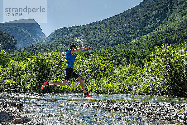 Männlicher Trailrunner springt an einem sonnigen Tag in den Bergen über einen felsigen Fluss