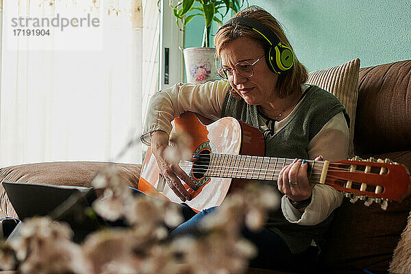 Frau mittleren Alters mit Kopfhörern spielt Gitarre auf dem Sofa zu Hause