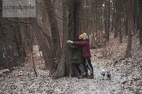 Lesbische Paar umarmen einen Baum zusammen mit kleinen Hunden im Winter Deutschland