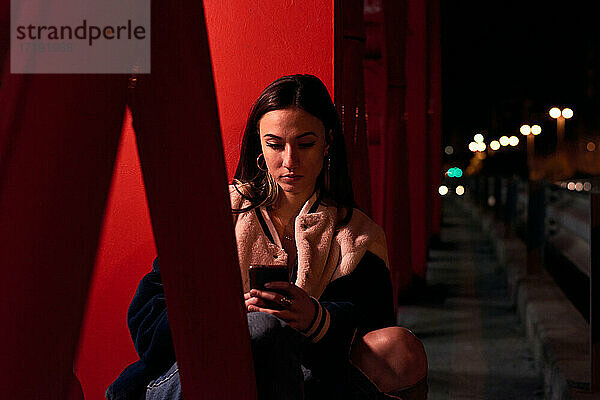 Junge Frau sitzt und schaut auf ihr Telefon auf einer roten Brücke