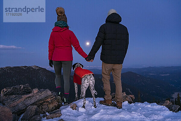 Rückansicht eines Paares  das sich an den Händen hält  während es mit seinem Hund auf einem Berggipfel in der Dämmerung steht
