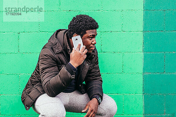 Junger schwarzer afroamerikanischer Junge  der sich hinhockt und mit seinem Mobiltelefon auf grünem Wandhintergrund spricht.