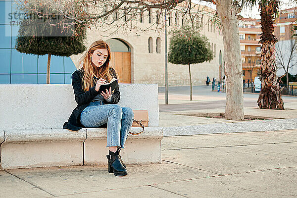 Junge Frau schreibt auf einer Steinbank sitzend in ihr Notizbuch