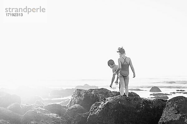 Junge und Mädchen  die bei Ebbe und Sonnenuntergang auf den Felsen am Strand spazieren gehen.