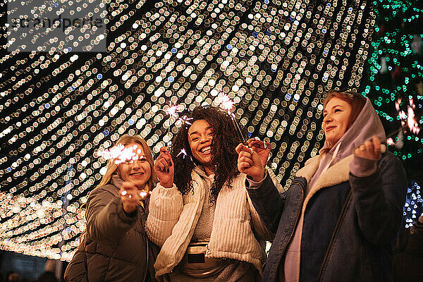 Glückliche Freundinnen  die Wunderkerzen in der Hand halten und vor einer Weihnachtsbeleuchtung stehen