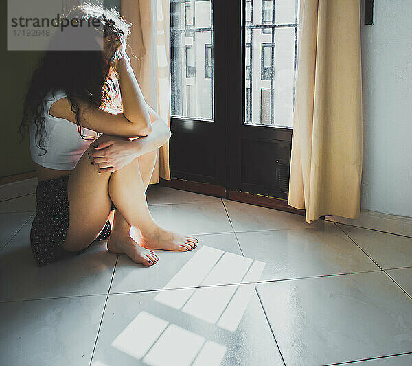 junge Frau  die besorgt auf dem Boden sitzt und aus dem Fenster schaut