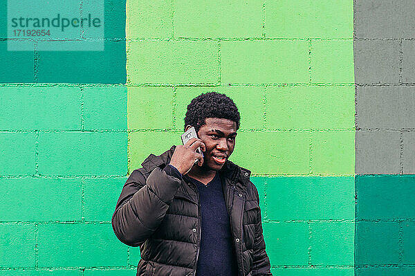Junger schwarzer afroamerikanischer Junge im Gespräch mit seinem Mobiltelefon auf grünem Wandhintergrund.