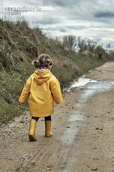 ein zweijähriges Mädchen in einem gelben Regenmantel  das in eine Schlammpfütze läuft