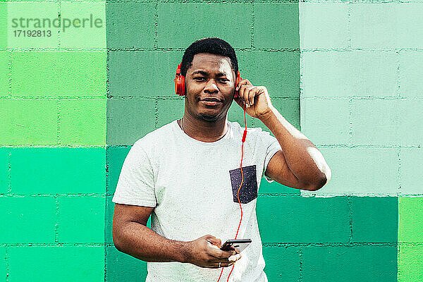 Afroamerikanischer schwarzer Junge auf grünem Wandhintergrund. Hören von Musik mit Kopfhörern und Mobiltelefon.