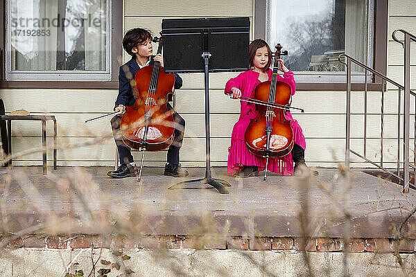 Zwei Kinder sitzen in Kostümen draußen und spielen Cello auf der Veranda