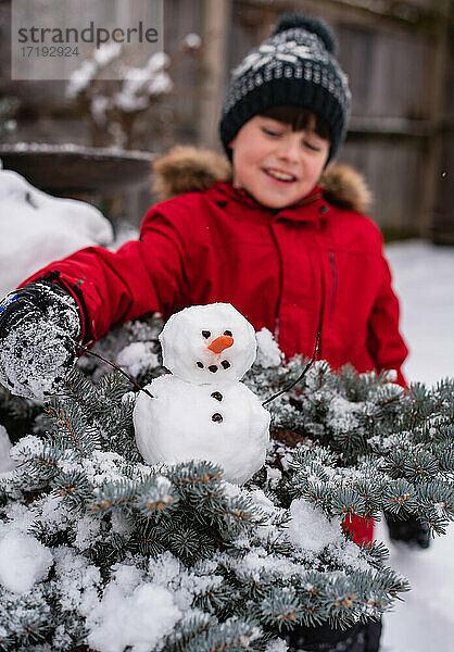 Junge betrachtet einen kleinen Schneemann auf einem Strauch im Freien an einem verschneiten Tag.
