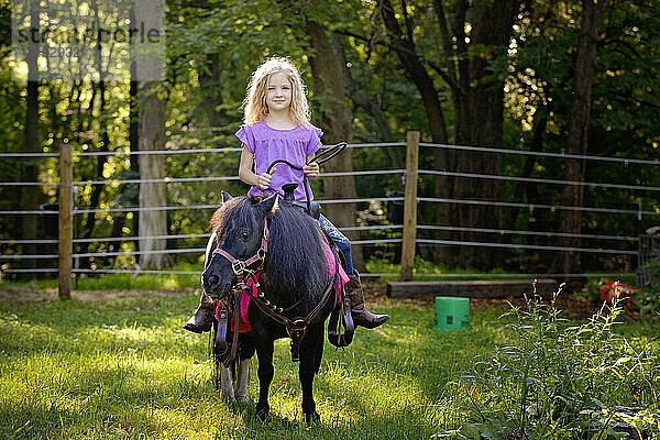 Schönes blondes Mädchen in lila Shirt reitet auf einem kleinen schwarzen Pony.