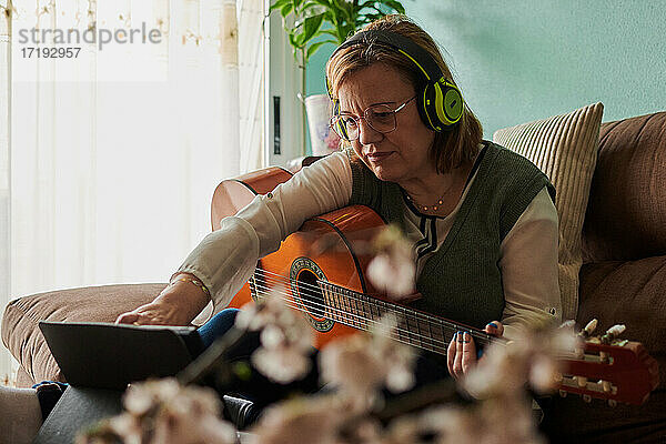 Frau mittleren Alters mit Kopfhörern spielt Gitarre auf dem Sofa zu Hause