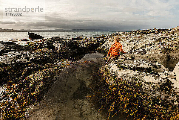 Kind in orangefarbener Jacke schaut vom Gezeitenbecken in Neuseeland auf den Ozean