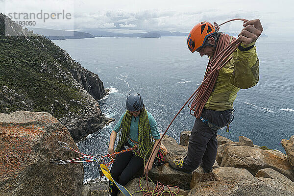 Ein abenteuerlustiges Paar lacht beim Aufrollen des Seils  während es an einem bewölkten Tag die exponierten Klippen von Cape Raoul in Tasmanien  Australien  erklimmt.