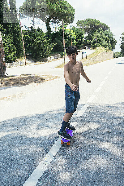 Porträt eines fröhlichen Teenagers ohne Hemd  der auf einer Bergstraße Skateboard fährt