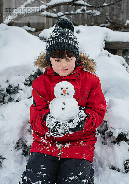 Junge hält an einem verschneiten Tag im Freien einen kleinen Schneemann in den Händen.