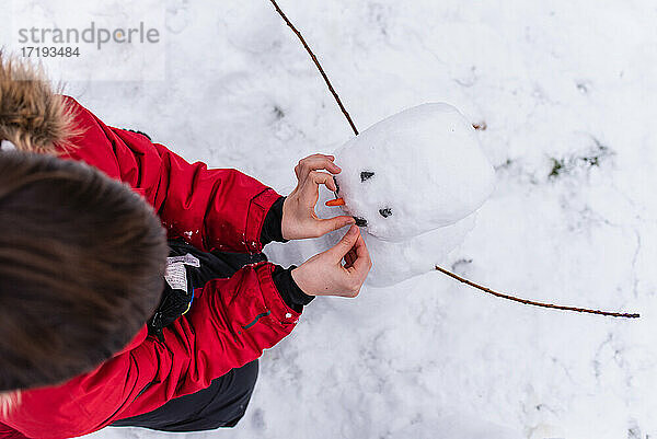 Draufsicht auf ein Kind im roten Mantel  das an einem Wintertag einen Schneemann baut.