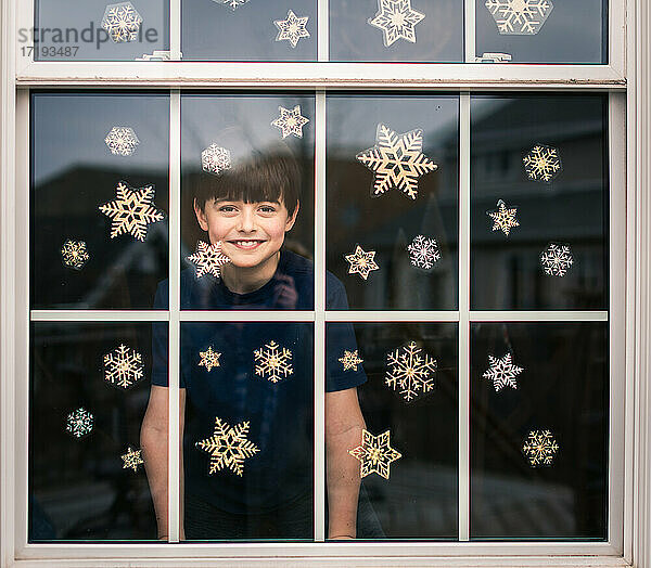 Fröhlicher Junge  der durch ein mit Schneeflocken bedecktes Fenster hinausschaut.