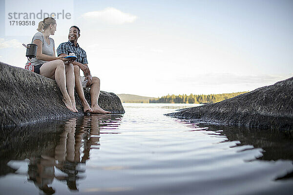 Mehrrassiges Paar sitzt auf einem Felsen am Rande eines Sees in Maine