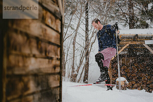 Ein Mann lacht im Schnee beim XC-Skifahren vor einer rustikalen Hütte