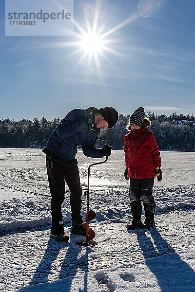 Zwei Jungen benutzen an einem sonnigen Wintertag einen Erdbohrer  um ein Loch ins Eis zu bohren.