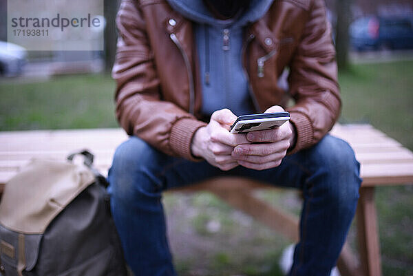 Das Bild konzentriert sich auf die Hände eines Mannes mit seinem Mobiltelefon auf einer Holzbank in einem Park.