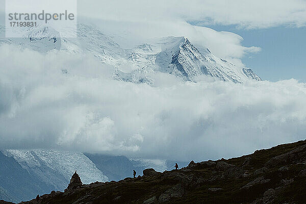 Zwei Wanderer wandern in der Ferne zwischen Wolken und verschneiten Gipfeln