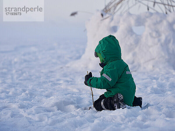 Kleiner Junge spielt mit Stock im Schnee an einem kalten Wintertag