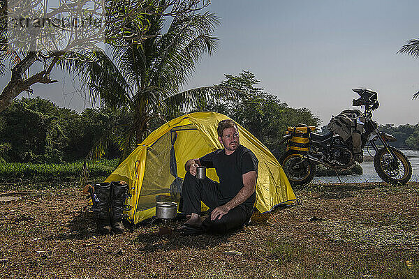 Mann zeltet mit seinem Geländemotorrad am Fluss Kwai