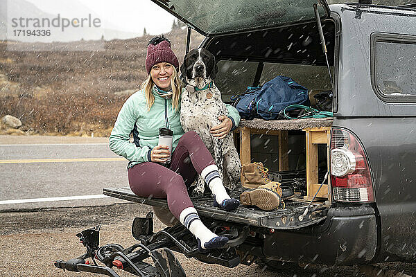 Lächelnde junge Frau sitzt mit Hund im Kofferraum eines Geländewagens bei Schneefall