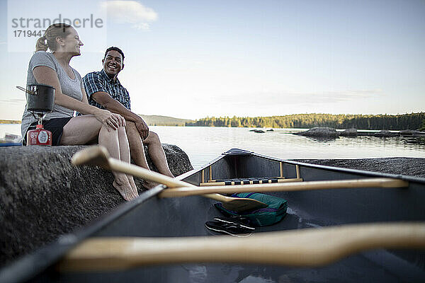Afroamerikanischer Mann und weiße Frau sitzen auf einem Felsen am See mit Kanu
