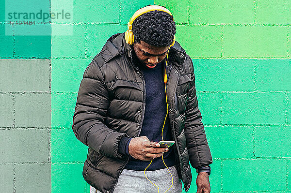 Schwarzer afroamerikanischer Junge auf grüner Hintergrundwand. Hören von Musik mit gelben Kopfhörern und sein Mobiltelefon.