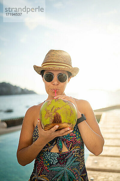 Glückliche Frau  die sich im Schwimmbad entspannt und Kokosnusswasser trinkt.