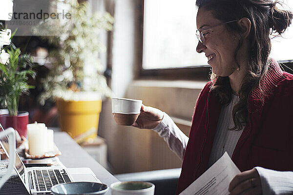 Frau lächelt mit einer Tasse Tee bei der Arbeit am Laptop im Home Office