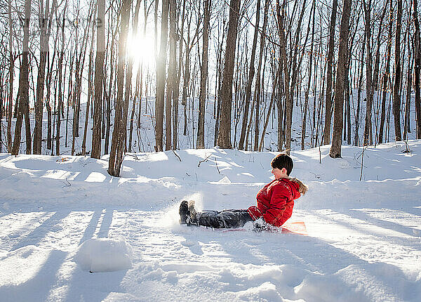 Junges Kind schlittelt einen verschneiten Hügel in einem Waldgebiet an einem sonnigen Tag hinunter.