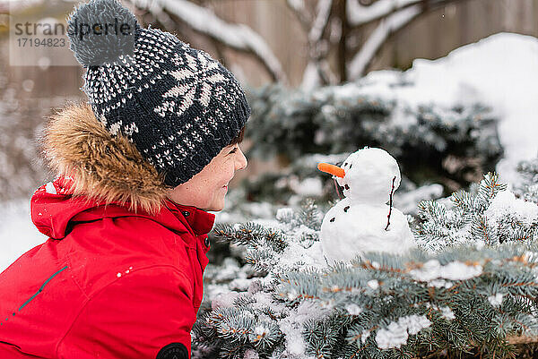 Junge betrachtet einen kleinen Schneemann auf einem Strauch im Freien an einem verschneiten Tag.