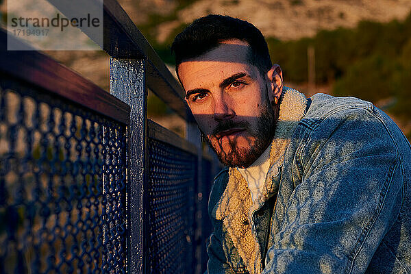 Hübscher Mann in Jacke posiert auf einer Brücke bei Sonnenuntergang