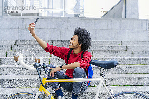 junger Mann mit Afro-Haar sitzt und macht ein Selfie neben seinem alten Fahrrad