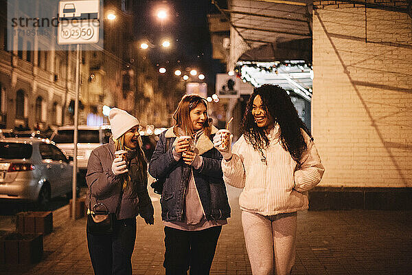 Glückliche weibliche Freunde mit heißen Getränken zu Fuß auf der Straße in der Stadt
