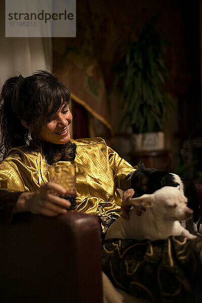 Frau lacht über kleine Hunde auf dem Schoß mit Getränk in der Hand beim Entspannen zu Hause