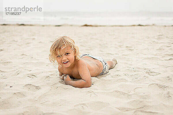 Blondes Kleinkind krabbelt auf dem Bauch in den Sand am Strand