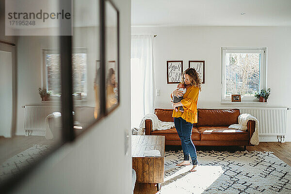 Mutter hält Baby im Wohnzimmer stehend mit Reflexion auf Bilder