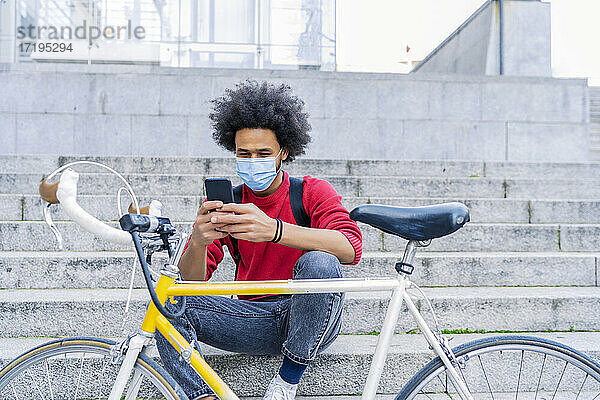 junger Mann mit Afrohaar  der neben seinem alten Fahrrad sitzt und eine Nachricht sendet
