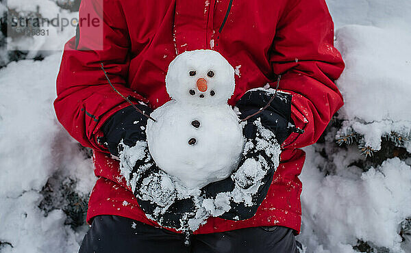 Kind hält an einem verschneiten Tag im Freien einen kleinen Schneemann in den Händen.