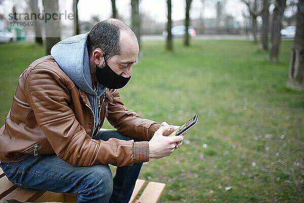 Kaukasischer Mann  der eine Maske trägt  um sich vor Covid19 zu schützen  schaut auf sein Handy auf einer hölzernen Parkbank  offener Raum.