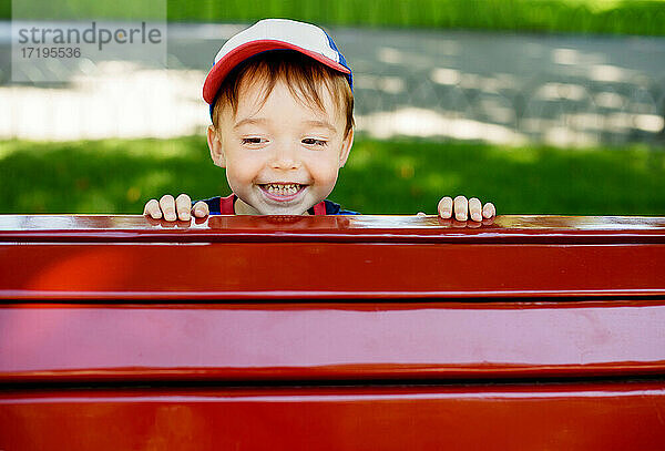 Cute Kleinkind lächelnd glücklich späht aus hinter roten Parkbank