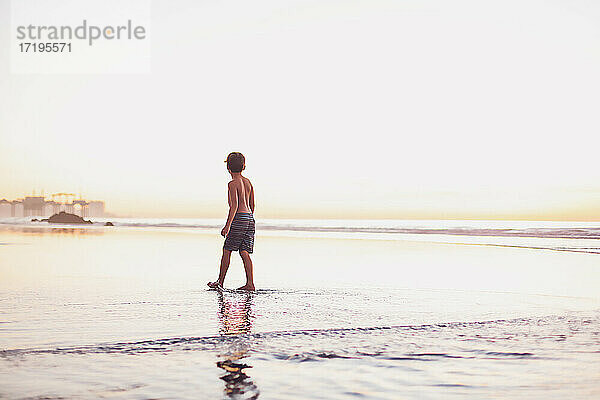 Junge  der während des Sonnenuntergangs bei Ebbe am Strand spazieren geht.