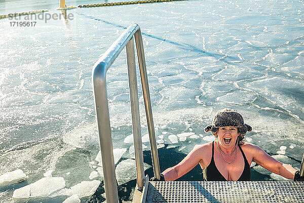 Amerikanische Frau mit Pelzmütze steigt über eine Leiter ins gefrorene Wasser in Dänemark hinab