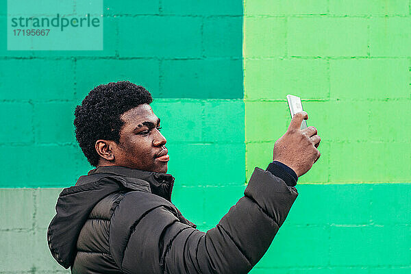 Junger schwarzer afroamerikanischer Junge  der ein Selfie mit seinem Mobiltelefon auf grünem Wandhintergrund macht.